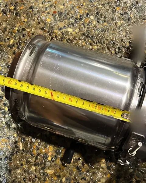 Cuerpo cilíndrico de acero inoxidable de un filtro de partículas, con una cinta métrica encima, sobre el suelo del taller.