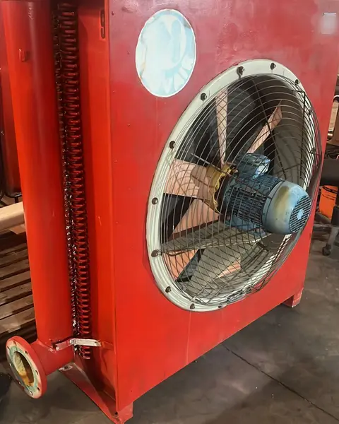 Intercambiador de calor industrial pintado en rojo, con un ventilador axial en la tapa y una brida de conexión abajo, colgado de cadenas en la nave.