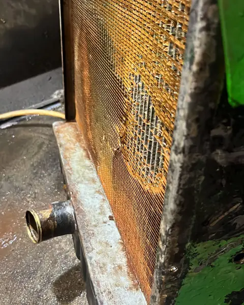 Radiador visto de lado, con el panal de cobre corroído y un hueco sin aletas, sobre el suelo del taller.